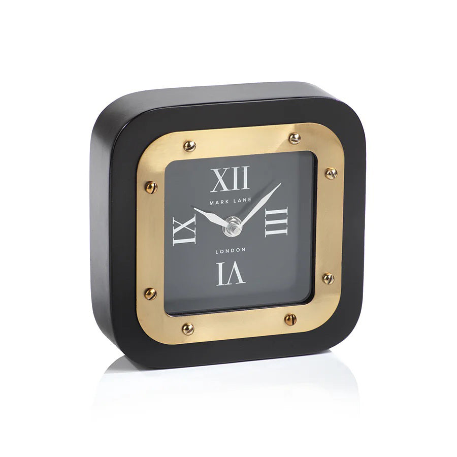 Two MARK LANE TABLE CLOCK - LARGE by ZODAX, square in shape with black and gold iron finish and Roman numerals, are displayed—one on a table, the other atop &quot;Rooms to Inspire&quot; books. The clocks show different times.