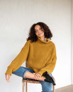 A woman with curly hair stands against a white wall, wearing a chunky mustard-colored MERSEA - BEACHSIDE TURTLENECK and blue jeans. She holds the hem of her turtleneck with one hand and has a relaxed expression.