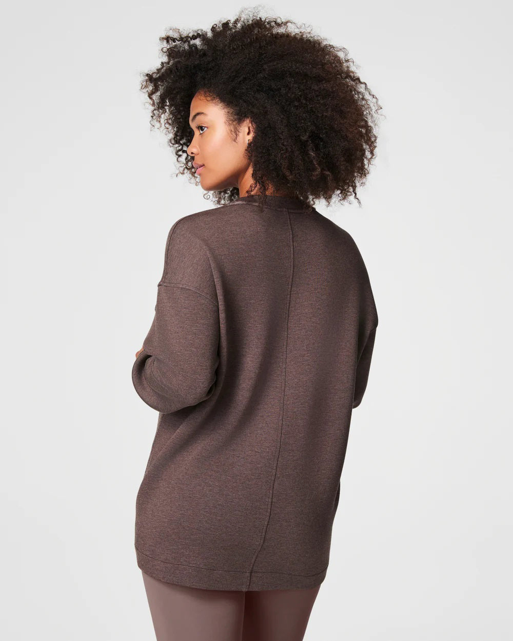 A person with curly hair is standing and looking to the side, wearing a cozy SPANX - AIRESSENTIALS BRUSHED V-NECK TUNIC paired with brown pants. The breathable top drapes elegantly, capturing a sense of laid-back comfort against the plain, light-colored background.