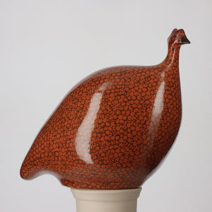 Introducing the LES CERAMIQUES DE LUSSAN - MEDIUM CERAMIC FRENCH GUINEA IN TERRACOTTA RED SPOTTED BLACK, a beautifully handmade ceramic sculpture from the South of France. This piece features a glossy red-orange finish with a distinctive black speckled pattern. The stylized, rounded form of the guinea fowl is perched elegantly on a light-colored cylindrical base, set against a plain, light gray backdrop.