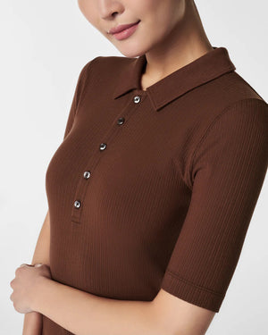 A woman in the SPANX - STRETCH RIB POLO TOP, a brown, ribbed, short-sleeve shirt with a button-down front, stands with her arms relaxed. The plain white background accentuates the top's stretchy ribbed fabric and body-hugging fit.