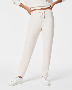 A person stands against a white background, smiling and looking down with one hand touching their hair. They are wearing a matching light beige SPANX AIRESSENTIALS TAPERED PANT and sweatshirt set made from ultra drapey, lightweight spacer fabric, paired with white sneakers.