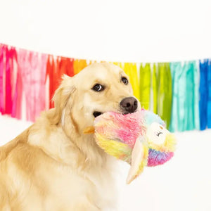 A golden retriever stands with the FRINGE STUDIO - BEE HOO YOU ARE DOG TOY by PET SHOP BY FRINGE STUDIO in its mouth, set against a background of colorful, hanging streamers arranged in a rainbow pattern. The toy features a super loud squeaker and crazy crinkle paper, adding an interactive play element to the vibrant scene.