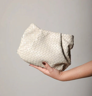 A person with painted nails holds the DANIELLA LEHAVI BALI MINI WOVEN BAG, an off-white clutch featuring handmade woven accents against a plain background. The bag showcases a textured, basket-like pattern and has a slouchy, unstructured shape.