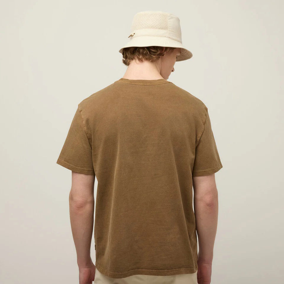 A young man in a beige bucket hat and a TILLEY Mid Weight Sunwashed T-Shirt in Peat stands against a light background, looking at the camera with a neutral expression.