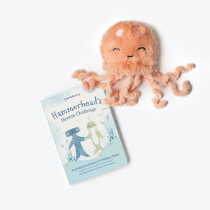 A children's book titled "Hammerhead's Recess Challenge" is displayed beside a plush jellyfish toy. The book features an illustration of a shark and a sea turtle on the cover and is subtitled "A Lesson in Conflict Resolution." The peach-colored jellyfish from SLUMBERKINS, perfect as a cuddle buddy, smiles cheerfully.