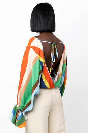 A person with shoulder-length black hair wears the HOPE FOR FLOWERS - BLOUSON TOP IN TUSCAN TILES, an off-shoulder blouse featuring rainbow-like horizontal stripes and made from Tencel. The blouse has flared sleeves adorned with similarly vibrant stripes and is paired with beige high-waisted pants. The individual poses against a plain white background, showcasing the colorful design of the top.