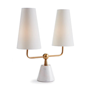 The MADISON DUBLET LAMP by NAPA HOME AND GARDEN showcases a sleek, contemporary design with a sophisticated white, conical marble base and two elegant white cylindrical lampshades. The lamp is supported by a gold metallic bar that gracefully splits into two upward arms featuring a brass finish, embodying modern elegance.