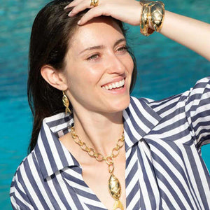 Close-up of a person wearing a pair of CAPUCINE DE WULF - POISSON DROP EARRINGS. The person's fingers, with light-colored nails, are touching their long dark hair. The image highlights the intricate design of the 18K Gold IP earring by CAPUCINE DE WULF and the person's ear and neck area.