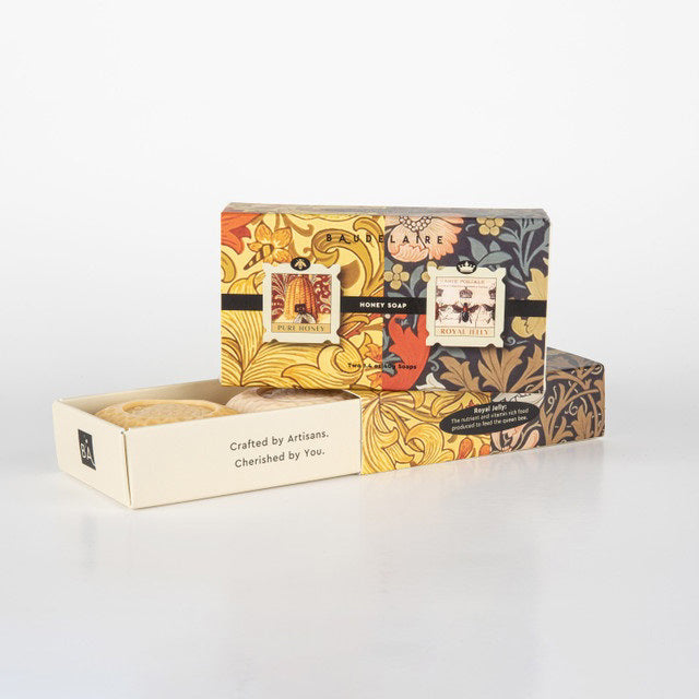 Two BAUDELAIRE soap boxes with floral designs, one opened to reveal a honeycomb patterned soap bar inside. Labels show &#39;Pure Honey&#39; and &#39;Royal Jelly&#39;. Text on the side reads &quot;Crafted by Artisans. Cherished by You.&quot; The BAUDELAIRE - PURE HONEY SOAP 1.4OZ MATCHBOX DUO is the perfect gift for any occasion. Background is plain white.