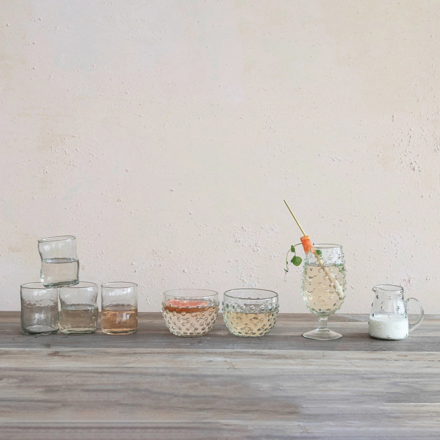 A variety of transparent drinking glasses, featuring a RECYCLED GLASS HOBNAIL STEMMED GLASS by CREATIVE COOP, is arranged on a wooden table. Some are stacked and filled with drinks. One has a garnished cocktail with a straw, and to the right, there's a small milk pitcher against a light beige wall.