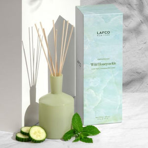 The LAFCO - Wild Honeysuckle Diffuser set features a green, angular glass bottle with wooden reeds inserted. The accompanying packaging, placed next to the bottle, showcases a light green and white marbled design with the LAFCO logo and scent description, accentuating the floral fragrance notes of this essential oil-based creation.