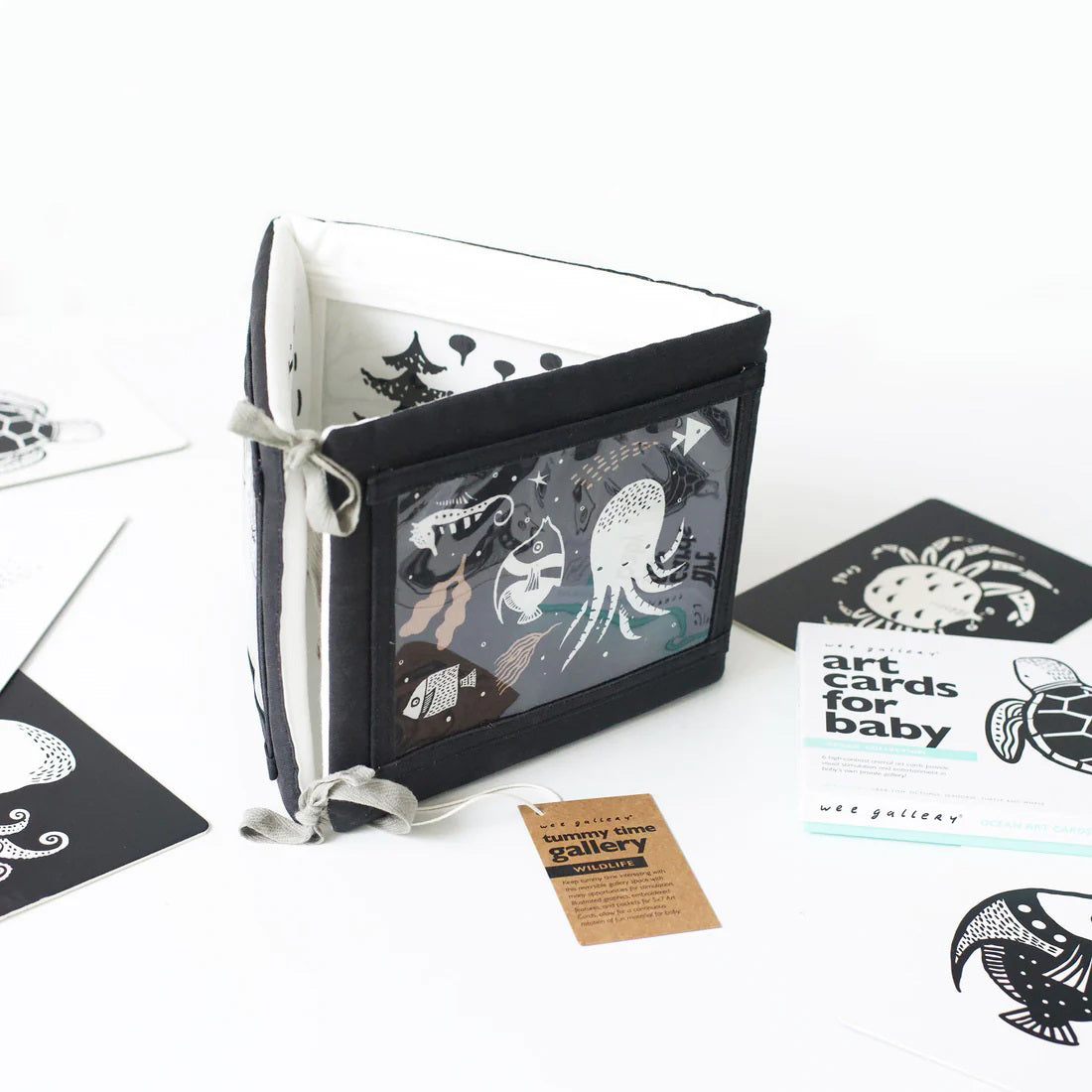 A TUMMY TIME GALLERY soft book from WEE GALLERY, featuring black and white fish and squid illustrations, is partially inserted in a clear front pocket. A brown tag attached by a string reads &quot;tummy time gallery.&quot; This Montessori toy, ideal for sensory development, rests on a plain white surface.