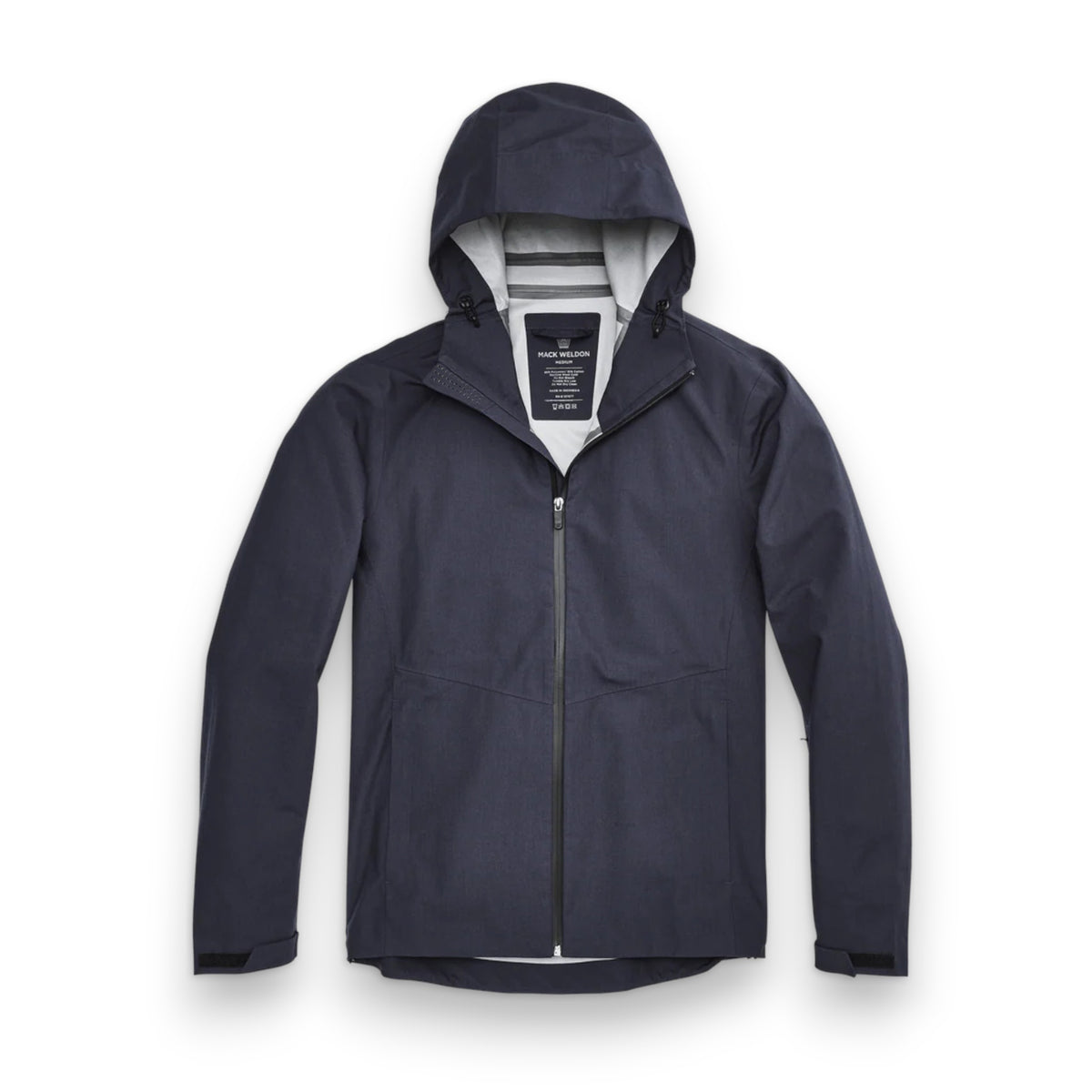 A man stands in front of a white wall wearing the MACK WELDON STORMCHASER JACKET with adjustable hood, paired with a gray shirt, blue pants, and black sneakers. He looks to the side while holding the jacket&#39;s zipper.