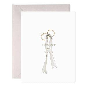 The WEDDING RINGS GREETING CARD by E.FRANCES PAPER showcases two gold rings tied with a gray ribbon on a white background, highlighted by shimmering foil accents. The phrase "FOREVER AND EVER" is elegantly printed in gold, and a beige envelope complements the card’s sophisticated charm.