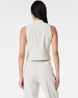 A person with long, straight hair is wearing the SPANX CAREFREE CREPE VEST, a light beige, sleeveless, tailored vest made of easy-care crepe fabric with three buttons and matching trousers from SPANX. Their hands are in their pockets as they pose in front of a plain white background.