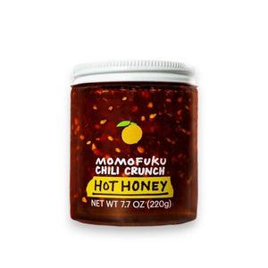 A jar of MOMOFUKU - CHILI CRUNCH HOT HONEY, with a white lid, contains a red-brown mixture inside. The label features an illustration of a citrus fruit along with the text "Momofuku Chili Crunch Hot Honey" and "Net Wt 7.7 oz (220g)." This spicy-crunchy chili oil offers the perfect blend of heat and sweetness.