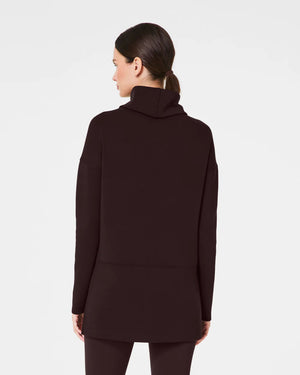 Dressed in the luxurious comfort of a SPANX AireEssentials Turtleneck Tunic, characterized by its black hue and lightweight fabric, a person stands with their back to the camera. Their elegantly tied up hair complements the matching black pants against a plain white backdrop.