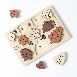 The WEE GALLERY - COUNT TO 10 WOODEN TRAY PUZZLE WITH LEAVES by WEE GALLERY is a wooden puzzle that includes ten oak leaf-shaped pieces in various shades of brown. Each piece fits into a light-colored wooden board adorned with minimalist drawings of trees and a bird. This educational toy is designed to enhance counting skills, featuring the text "Count to Ten" on the board.