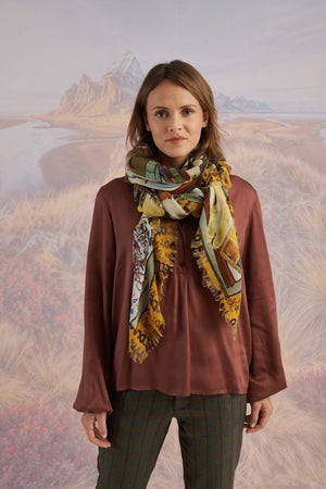 A woman with wavy blonde hair smiles while looking down. She wears a loose, light lavender blouse, dark pants, and the STORIATIPIC EUGENIE WOOL SCARF. Behind her is a scenic landscape of mountains, grass, and water.