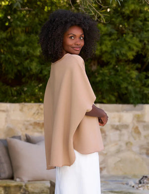 A woman with voluminous curly hair walks outdoors. She is wearing a loose-fitting FRANK AND EILEEN - OLIVE ORIGINAL CAPELET and white trousers, reminiscent of an original capelet. Behind her are lush green trees and a stone wall with a cushioned bench. She has a calm and confident expression.