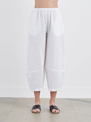 CUT LOOSE - LANTERN PANT by CUT LOOSE are white, wide-leg linen pants with an elastic waistband, hanging from a wooden hanger on a neutral-colored wall. These lightweight, breathable pants have a relaxed fit and feature visible seams at the knees.