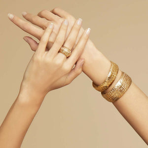 Two hands showcasing golden jewelry. The left hand sports a gold-plated woven ring, while the right hand is adorned with three thick, intricately designed gold bangles and the stunning GAS BIJOUX - WILD GOLD BRACELET by GAS BIJOUX. Both hands are positioned with elegance against a neutral background.