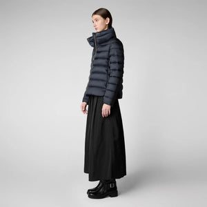 A person with dark hair is standing against a plain background, wearing a high neck SAVE THE DUCK - ELSIE PUFFER JACKET, a long black skirt, and black boots. They have a neutral expression and their arms are relaxed by their sides.