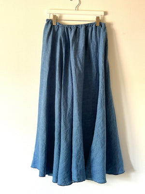 A flowy, long CP SHADES - DEIDRA SKIRT IN BLEACHED INDIGO TWILL hangs on a white hanger against a plain light background. This denim blue skirt from the CP SHADES collection features an elastic waistband and a slightly wrinkled texture, draping effortlessly to give it a casual and comfortable look.