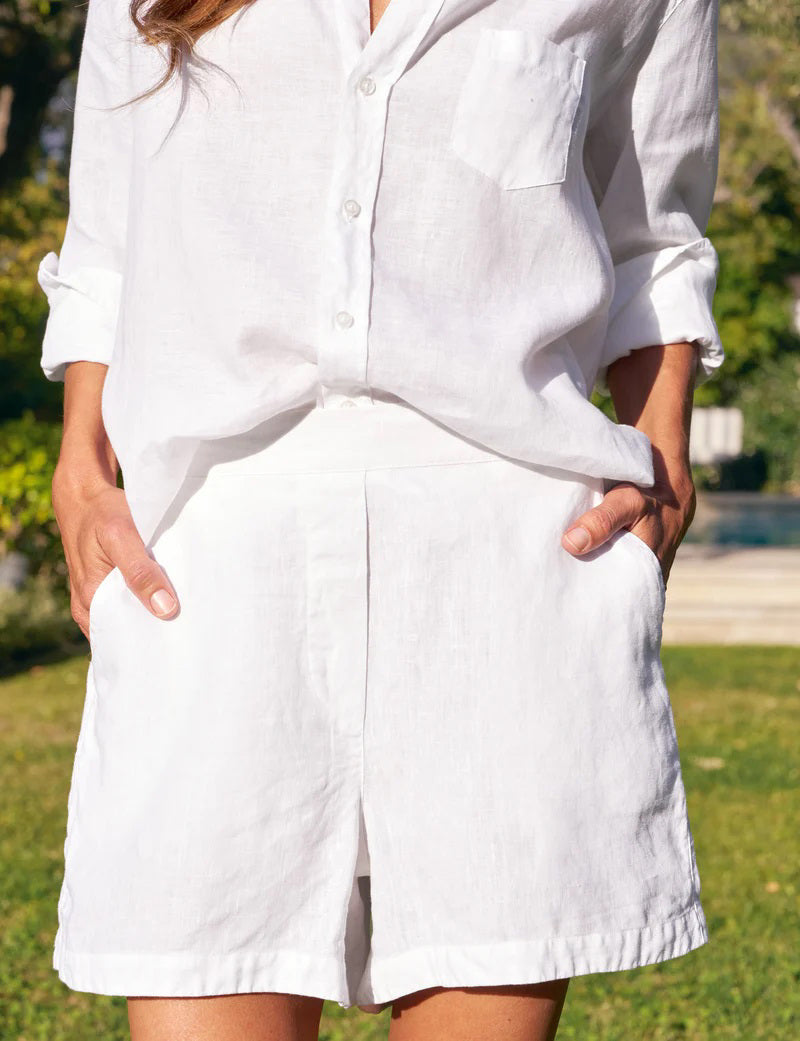 Wearing FRANK &amp; EILEEN&#39;s MAE LINEN SHORT, a person in a white button-up with rolled sleeves stands outdoors, hands in pockets; lush greenery and a pool behind evoke an effortless Amalfi Coast vibe.