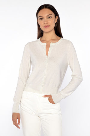 A person with long dark hair wears the KINROSS CASHMERE - SEQUIN DRESS CARDIGAN over a matching top and white pants, standing against a plain white background. The cardigan's subtle shimmer by KINROSS elegantly complements their neutral expression.