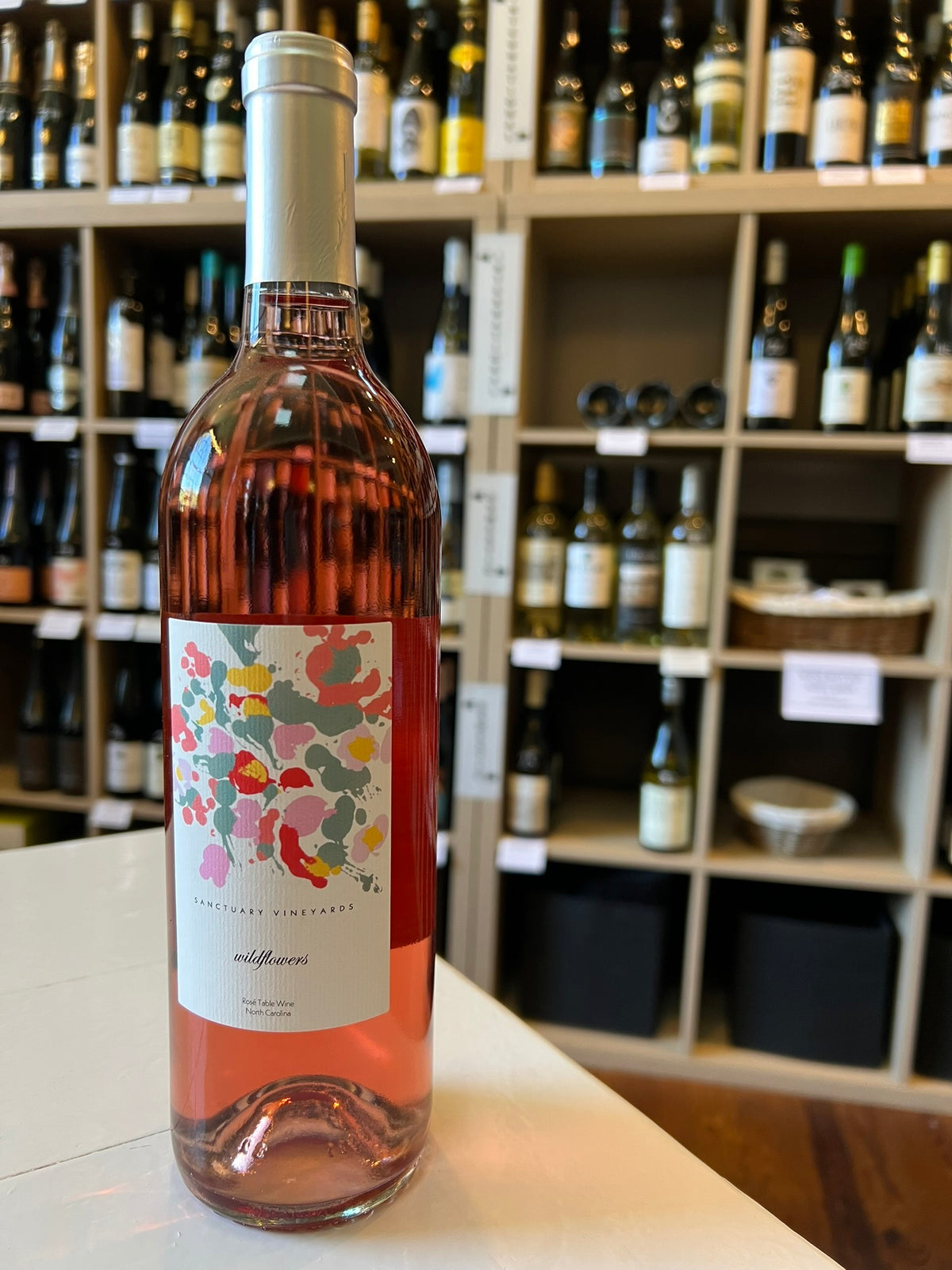 Enjoy a bottle of SANCTUARY &#39;WILDFLOWERS&#39; ROSÉ by WINE: FREEDOM BEVERAGE—light pink with a white label featuring colorful abstract flowers and a gold foil cap, perfect for experiencing North Carolina&#39;s unique wine flavors.