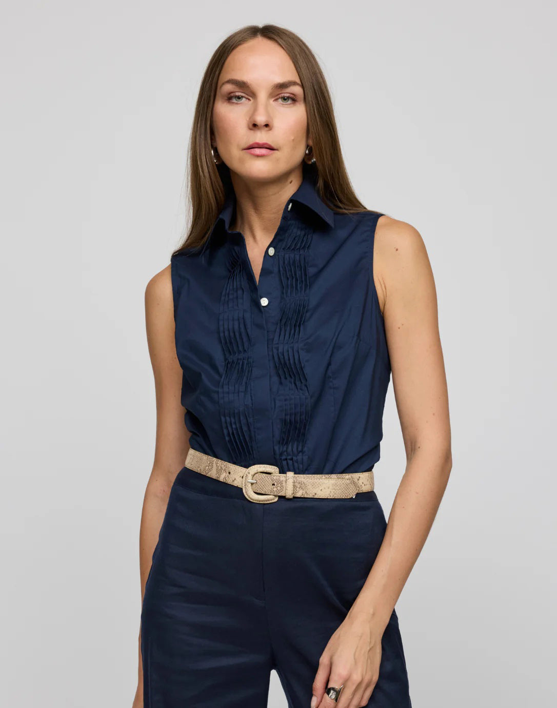 A woman with long hair wears the HINSON WU Lilith Sleeveless Shirt, made from stretch polished cotton with pleats and a beige belt, against a gray background. She exudes confidence as she gazes at the camera, highlighting the shirt's versatility.