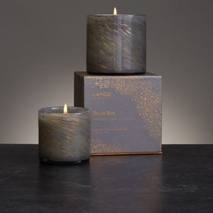 The LAFCO - FEU DE BOIS HOLIDAY CANDLE 6.5OZ is a stylish gray glass candle holder featuring a marbled texture, with a single white soy wax candle inside and delicate bronze swirls adorning the glass.