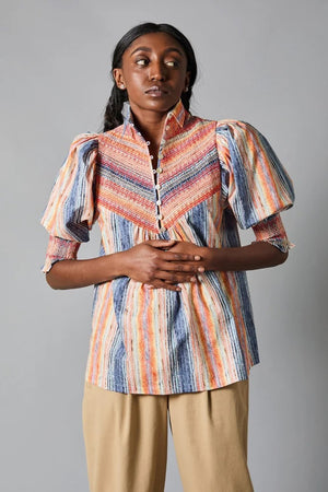 Against a gray backdrop, an individual with long, dark hair wears the HOPE FOR FLOWERS Smocked Shirt—an organic cotton blouse featuring colorful stripes, a smocked yoke, short puff sleeves, and a high collar—paired with beige pants. Their hands are clasped in front.