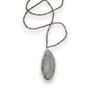 A unique jewelry piece, the ANN LIGHTFOOT Medium Agate Geode Pendant with Snowflake Jasper Strands features a large, polished oval agate geode slice pendant. The stone showcases a distinctive pattern with layered shades of gray, white, and beige that exudes an earthy and natural appeal. The necklace is beautifully displayed against a black background.