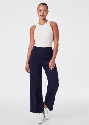 A person stands wearing a black short-sleeve top and the SPANX Stretch Twill Cropped Wide Leg Pant in brown, featuring CoreSure Tech for comfort. Paired with black sandals, they smile brightly, hands relaxed by their sides against a plain light gray background.