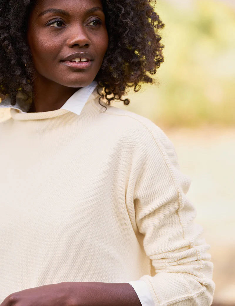 A woman with curly hair is wearing the luxurious FRANK & EILEEN - MONTEREY Rolled Funnel Neck Sweater in cream over a white collared shirt. She is looking to her right, and the background features outdoor greenery with a soft focus.