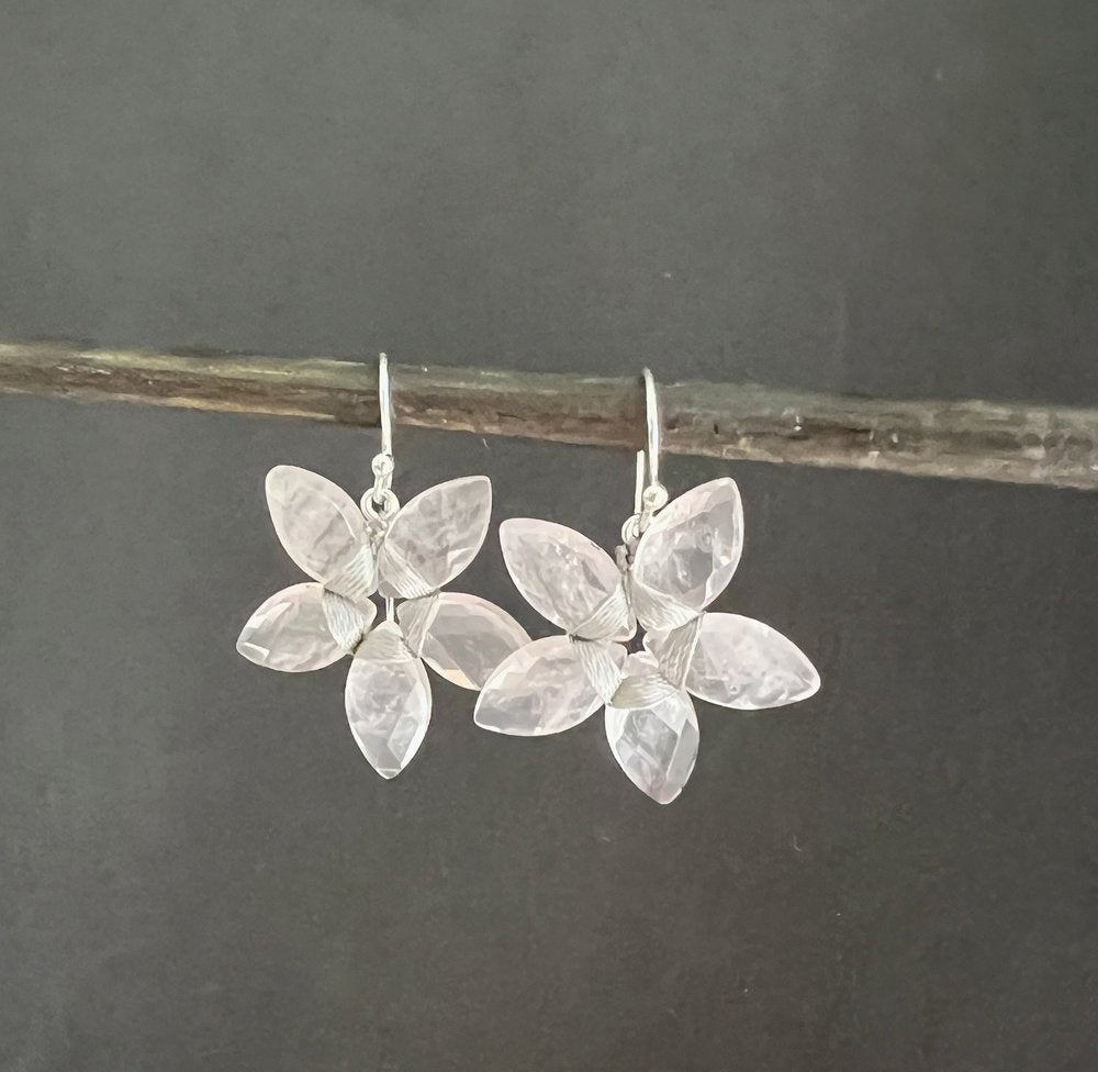 ANN LIGHTFOOT's Rose Quartz Marquise Cut Flower Earrings hang delicately from a thin branch against a dark backdrop, their translucent stones evoking rose quartz in bloom.