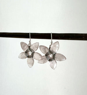 ANN LIGHTFOOT's Rose Quartz Marquise Cut Flower Earrings hang delicately from a thin branch against a dark backdrop, their translucent stones evoking rose quartz in bloom.