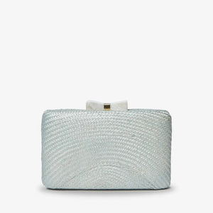 The KAYU - ELOISE CLUTCH, from brand KAYU, is a rectangular woven straw clutch with a light shimmer and a small white bow clasp. It can be styled with an optional chain strap, all set against a plain white background.