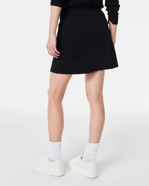 A person stands against a plain white background wearing a matching white long-sleeved sweater and the SPANX - AIRESSENTIALS SKORT by SPANX. Their hair is tied back in a low bun, and they are looking slightly to the side. They complete their look with white ankle socks and sneakers, highlighting the skort's hidden pocket feature perfectly.