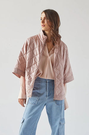 A person with shoulder-length hair stands facing sideways, wearing the ADROIT ATELIER - RUBY QUILTED CAPE, a light pink, short-sleeved jacket with a 100% Nylon shell, paired with light blue jeans. The background is plain white. The individual gazes off to the side, showcasing the back and side profile of the outfit from ADROIT ATELIER.