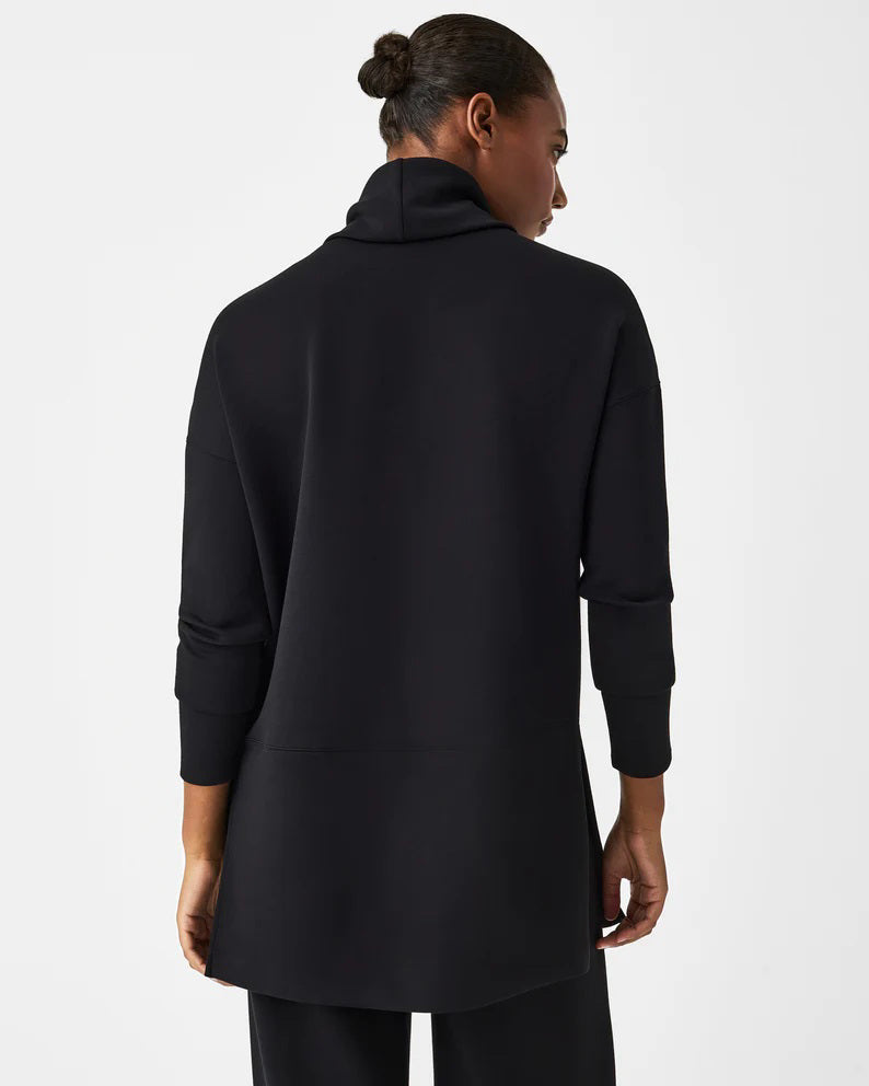 Dressed in the luxurious comfort of a SPANX AireEssentials Turtleneck Tunic, characterized by its black hue and lightweight fabric, a person stands with their back to the camera. Their elegantly tied up hair complements the matching black pants against a plain white backdrop.