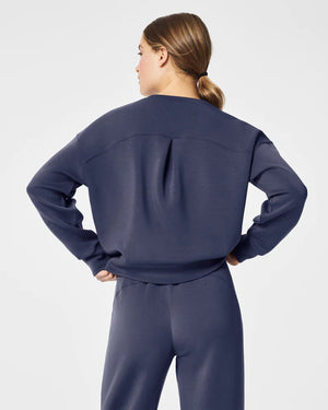 A woman stands confidently against a plain white background, wearing the SPANX Aireessentials Crew Neck Pullover and matching sweatpants made from luxurious comfort spacer fabric. She has her hair pulled back and her left hand is raised slightly. She smiles gently with her lips closed.