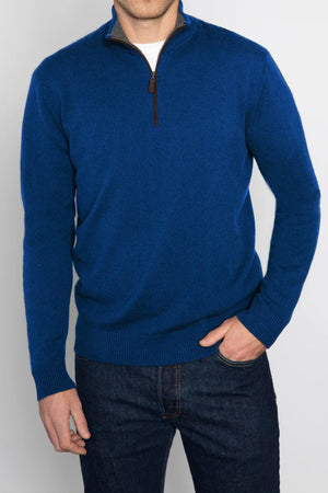 A person is wearing the KINROSS CASHMERE Men's Cashmere Suede Trim Quarter Zip Mock in blue over a white shirt and dark jeans. They have one hand in their pocket and stand against a plain, light-colored background. The quarter-zip sweater features a brown zipper pull.