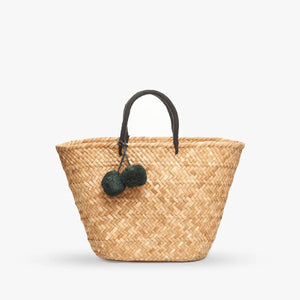 In a garden setting, a woman in a strapless beige top and white wrap skirt holds the KAYU Oversized St. Tropez Tote, adorned with black pom-pom accents, against a backdrop of lush grass and trees.