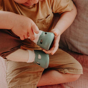 A close-up of a baby wearing OLLI ELLA USA DINKUM DOLL SHOES in Mallow Pink and white socks. The baby is seated on a soft, pale pink textured blanket, dressed in light brown clothing. Only the lower half of the baby's body is visible.