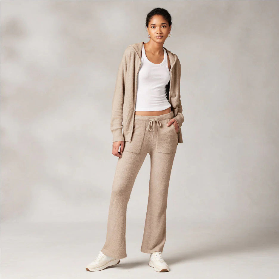 A woman is standing against a plain backdrop, wearing a beige hoodie made from Kash-ease yarn, KASHWERE's Wide Leg Lounge Pant with a matching drawstring waistband, a white tank top, and white sneakers. She has one hand in her pocket and the other by her side, looking directly at the camera.