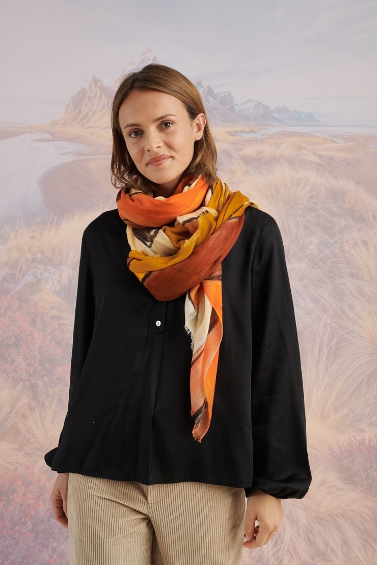 A woman in a black long-sleeve top and beige pants stands before a scenic backdrop, her look elevated by the STORIATIPIC ISCHIA Modal and Wool Scarf (100x200 cm) in vibrant orange and brown, draped elegantly around her neck.